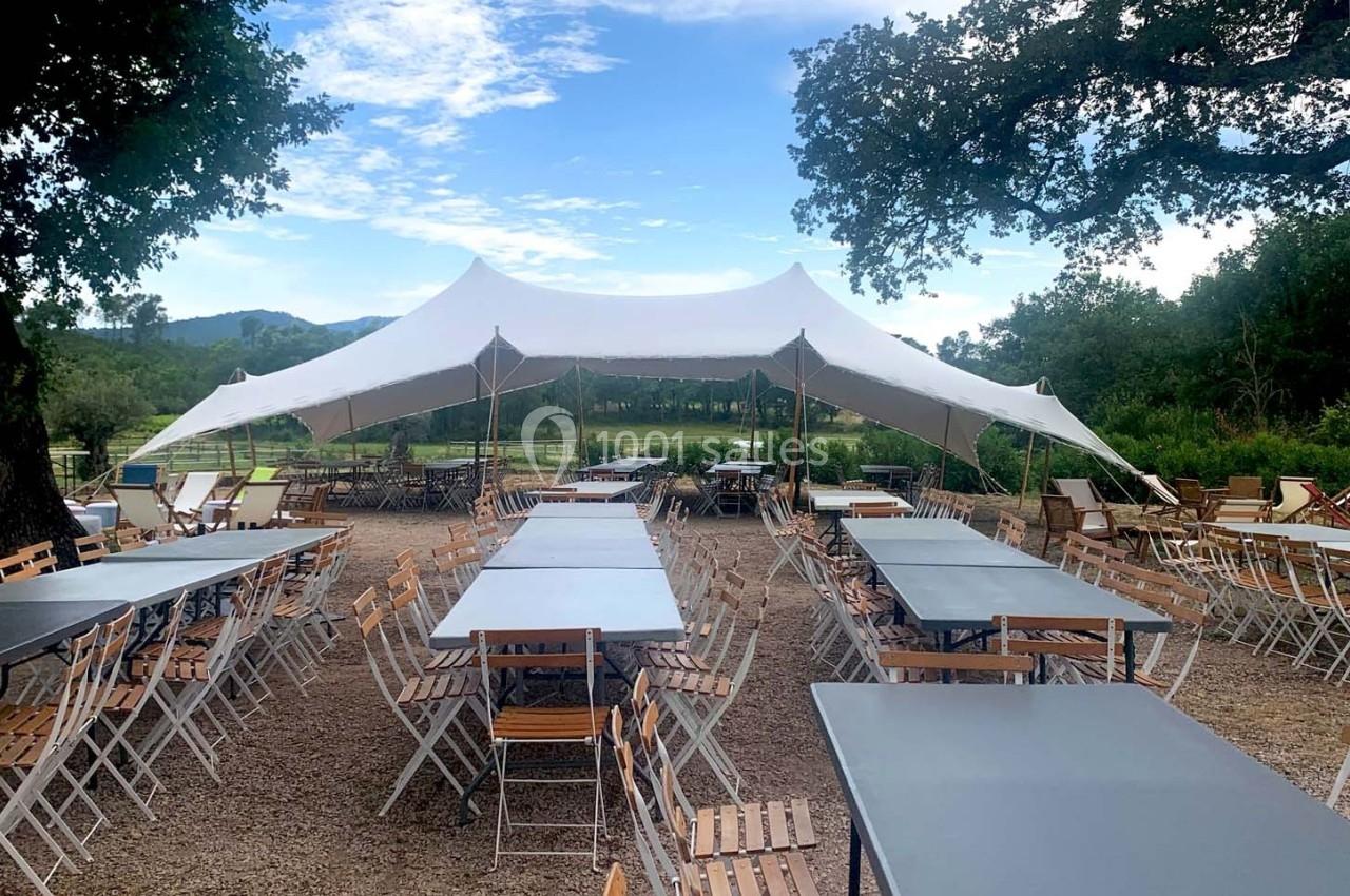 Tables et chaises en bois et métal disposées sous une grande tente blanche dans un espace extérieur arboré.