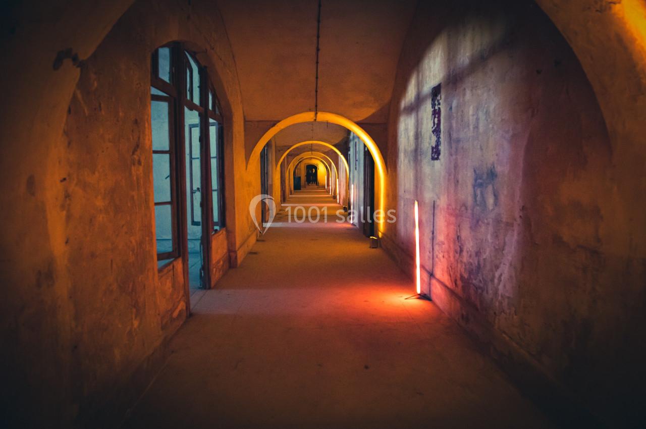 Couloir voûté éclairé par des lumières colorées, avec des murs usés et des fenêtres sur un côté.