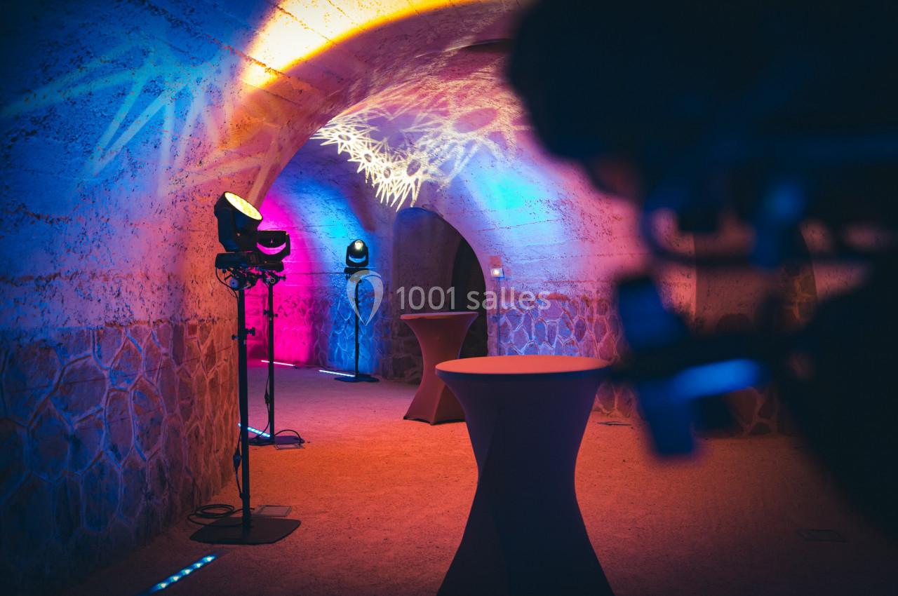 Salle voûtée éclairée par des lumières colorées, avec des tables hautes et des projecteurs disposés au sol.