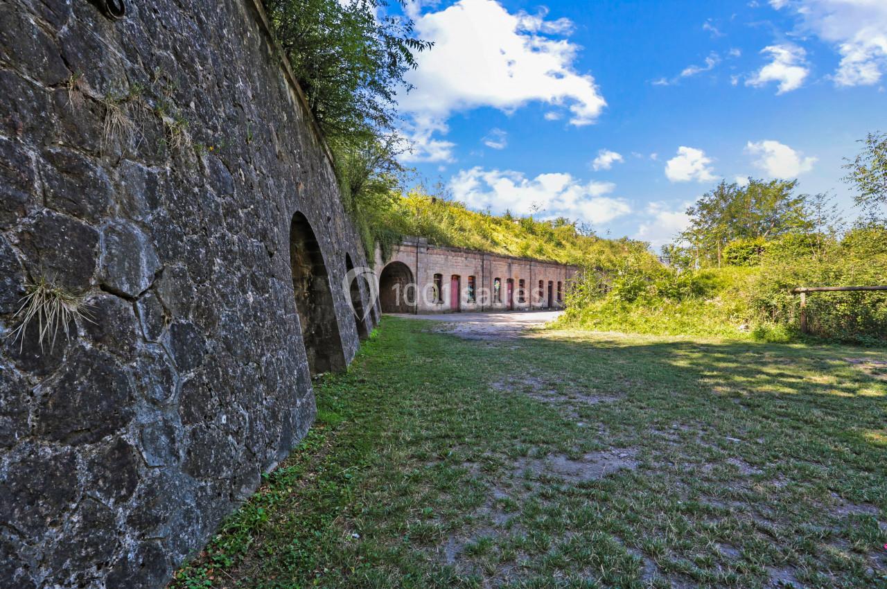 Mur en pierre d'un fort avec des arches, entouré de végétation et éclairé par un ciel bleu partiellement nuageux.