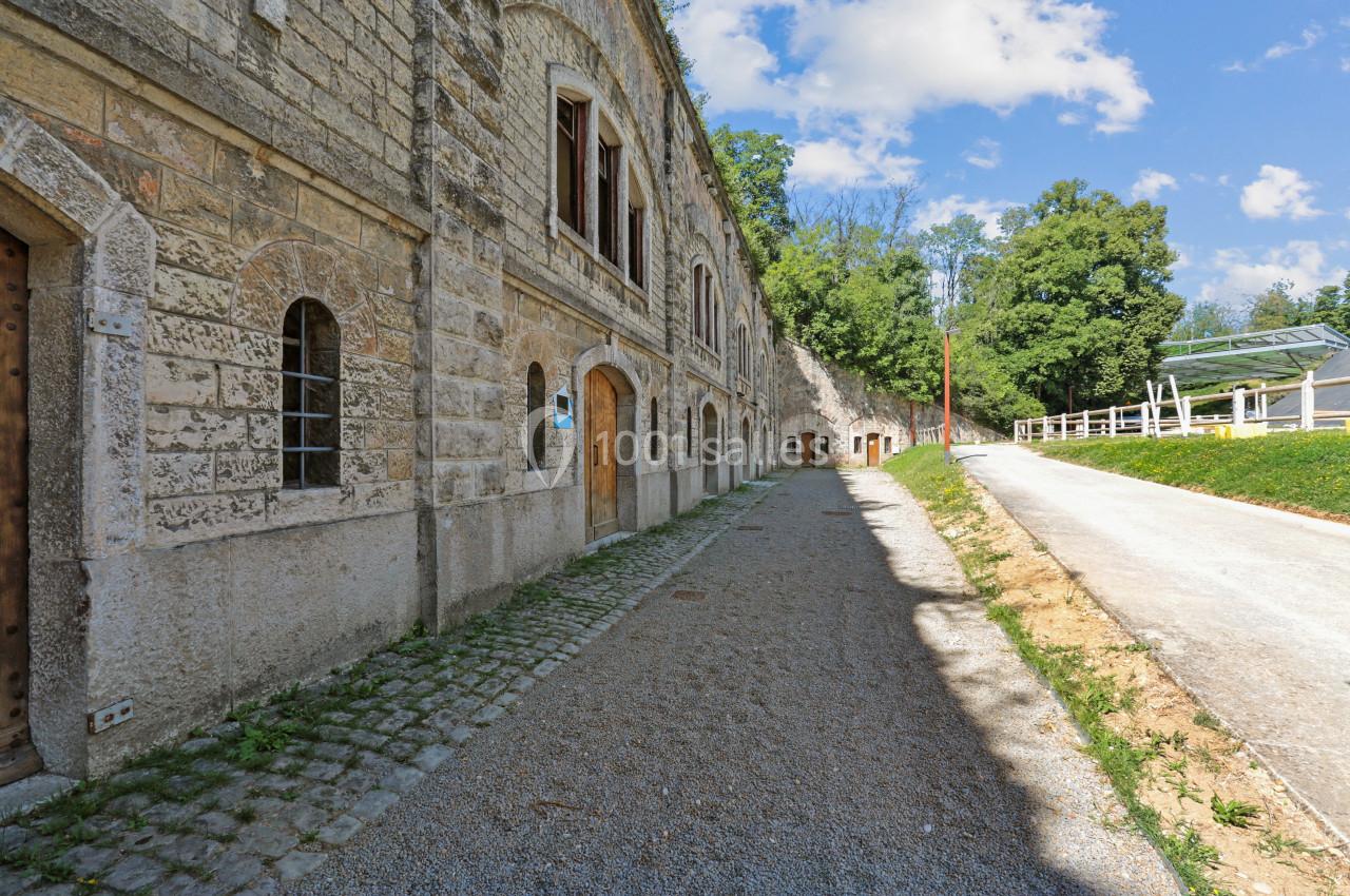 Façade en pierre d'un bâtiment historique longeant un chemin gravillonné, entouré de verdure et d'arbres.