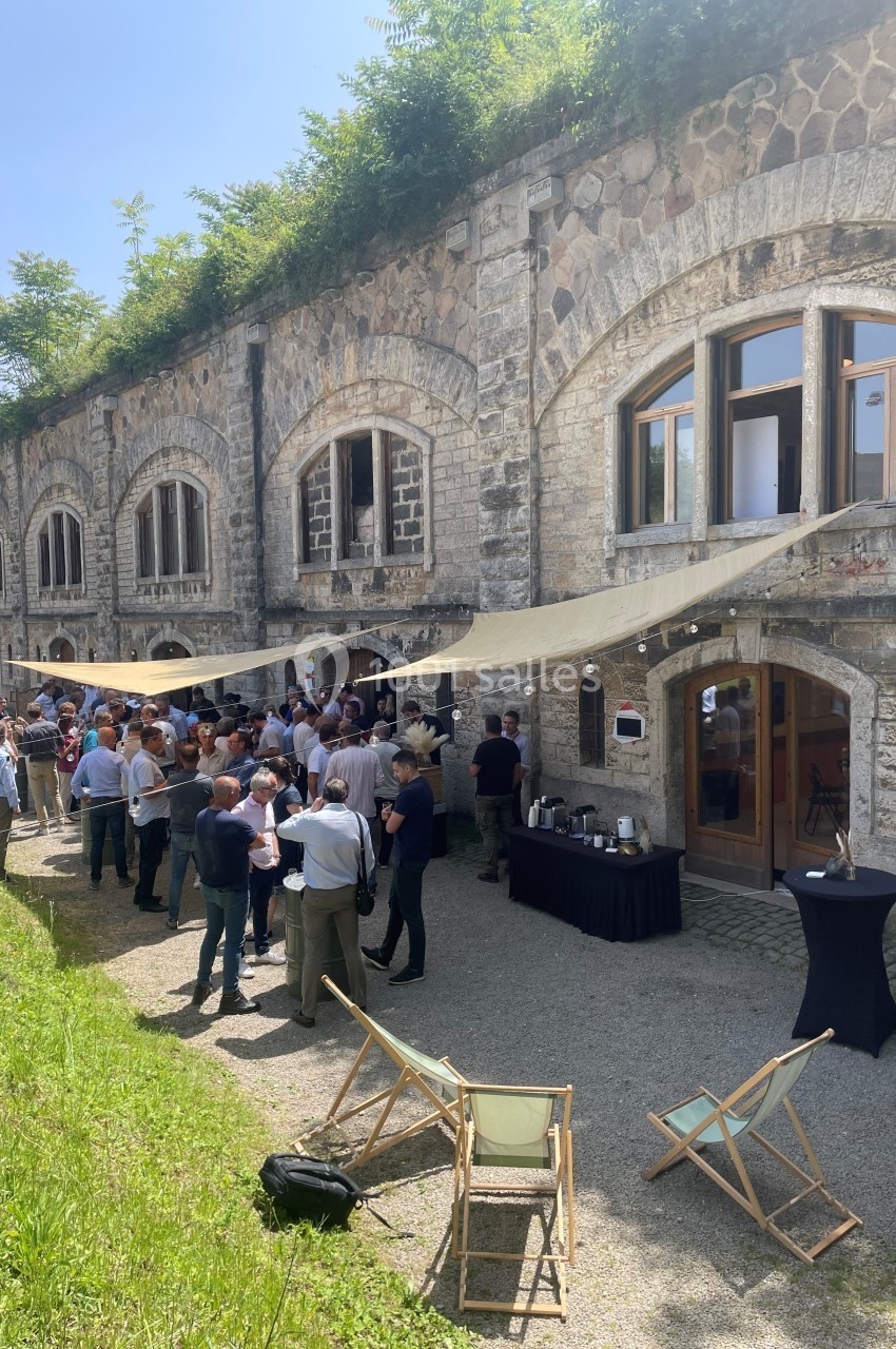 Groupe de personnes rassemblées devant un bâtiment en pierre, sous des toiles d'ombrage, avec des tables et chaises…
