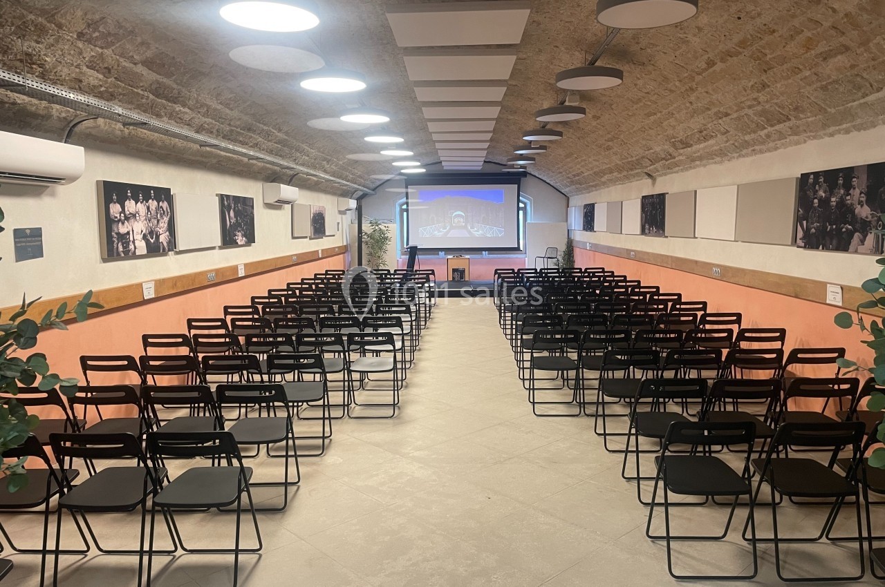 Salle voûtée aménagée avec des rangées de chaises noires face à un écran de projection allumé.