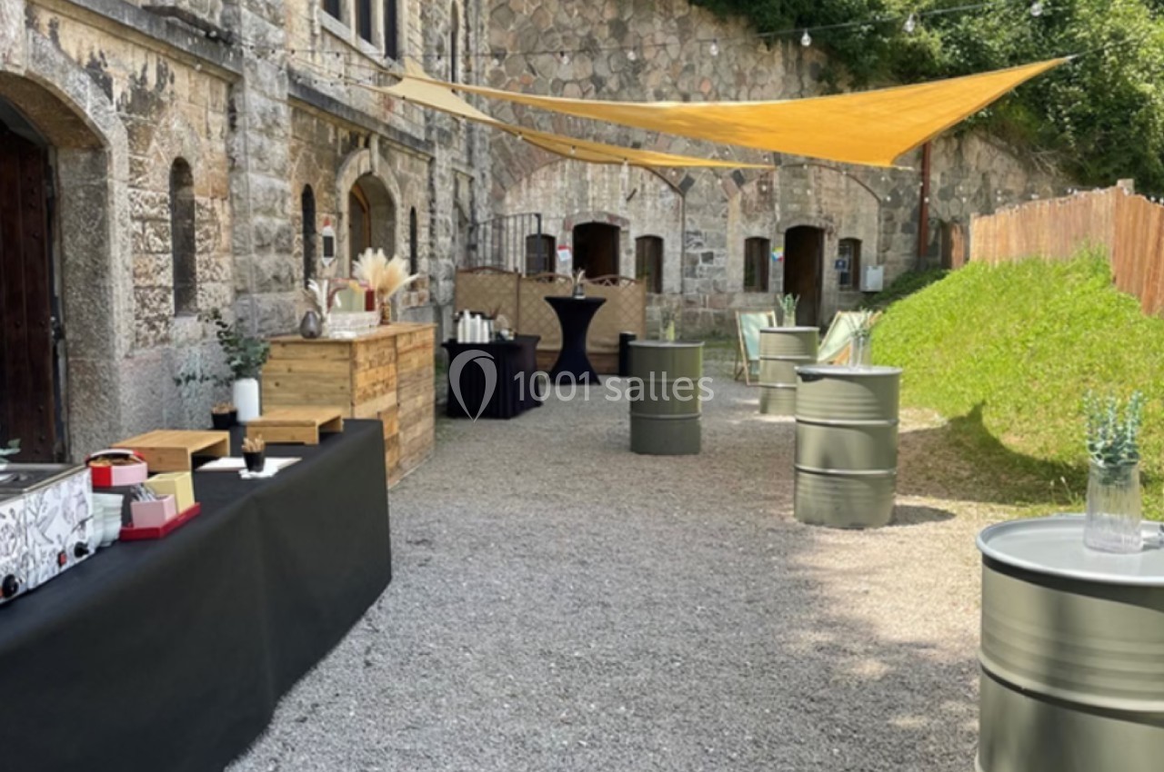 Cour extérieure avec des tables en tonneaux, des nappes noires et des voiles d'ombrage près d'un bâtiment en pierre.