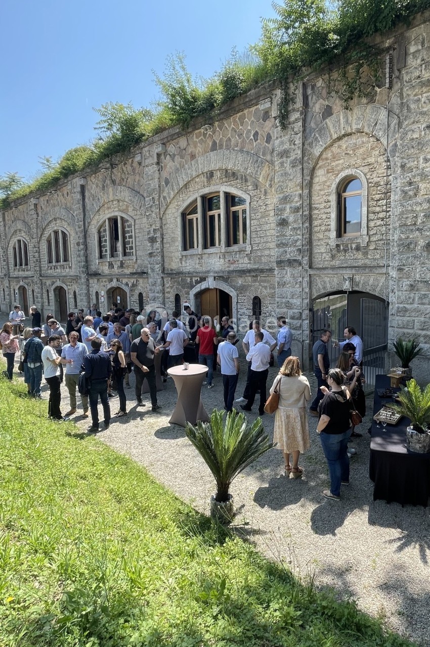 Groupe de personnes rassemblées devant un bâtiment en pierre avec des arches, dans un espace extérieur ensoleillé.
