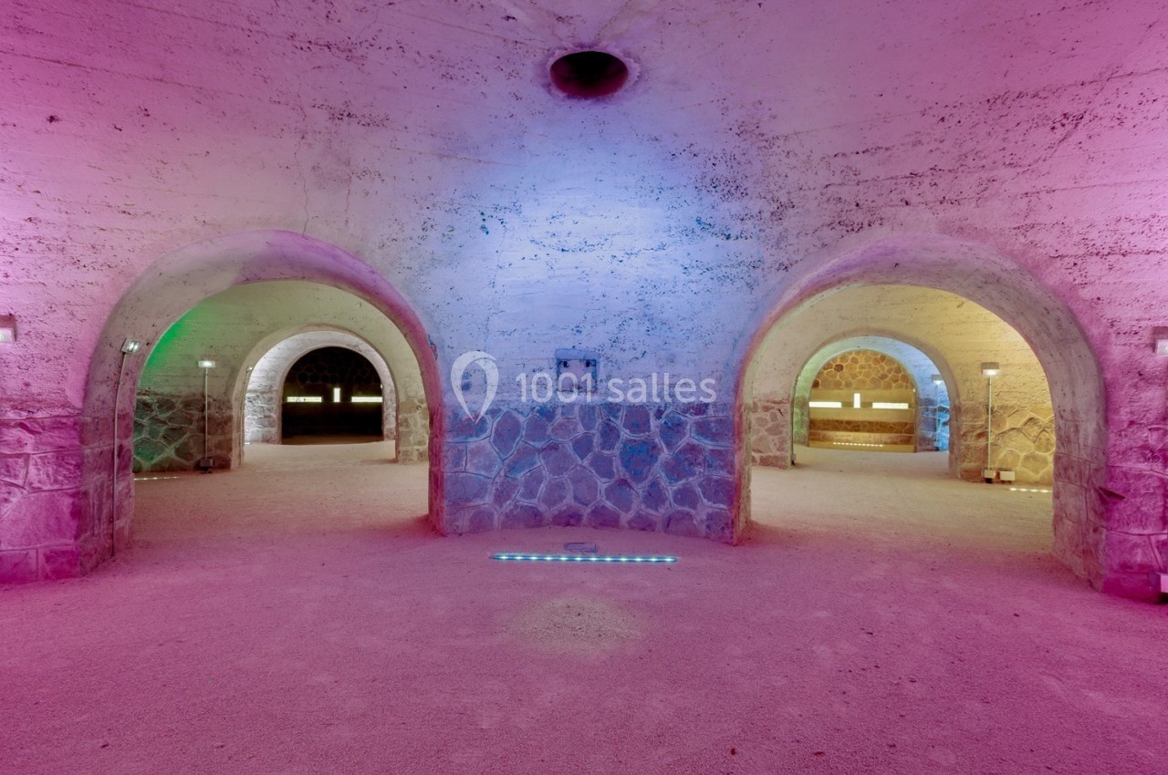 Salle voûtée en pierre avec éclairage coloré violet et bleu, trois arches menant à des espaces distincts.