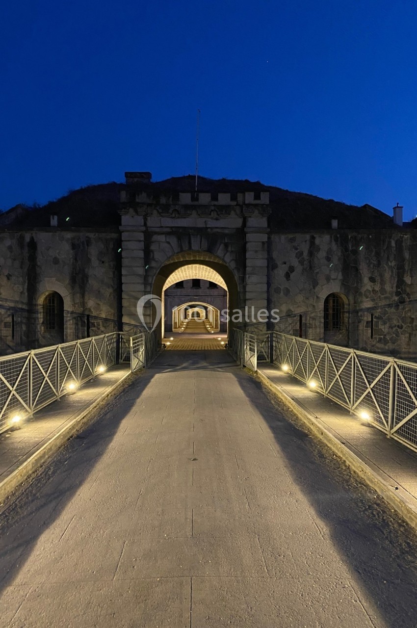 Passerelle éclairée menant à une entrée voûtée d'un fort en pierre, photographiée à la tombée de la nuit.