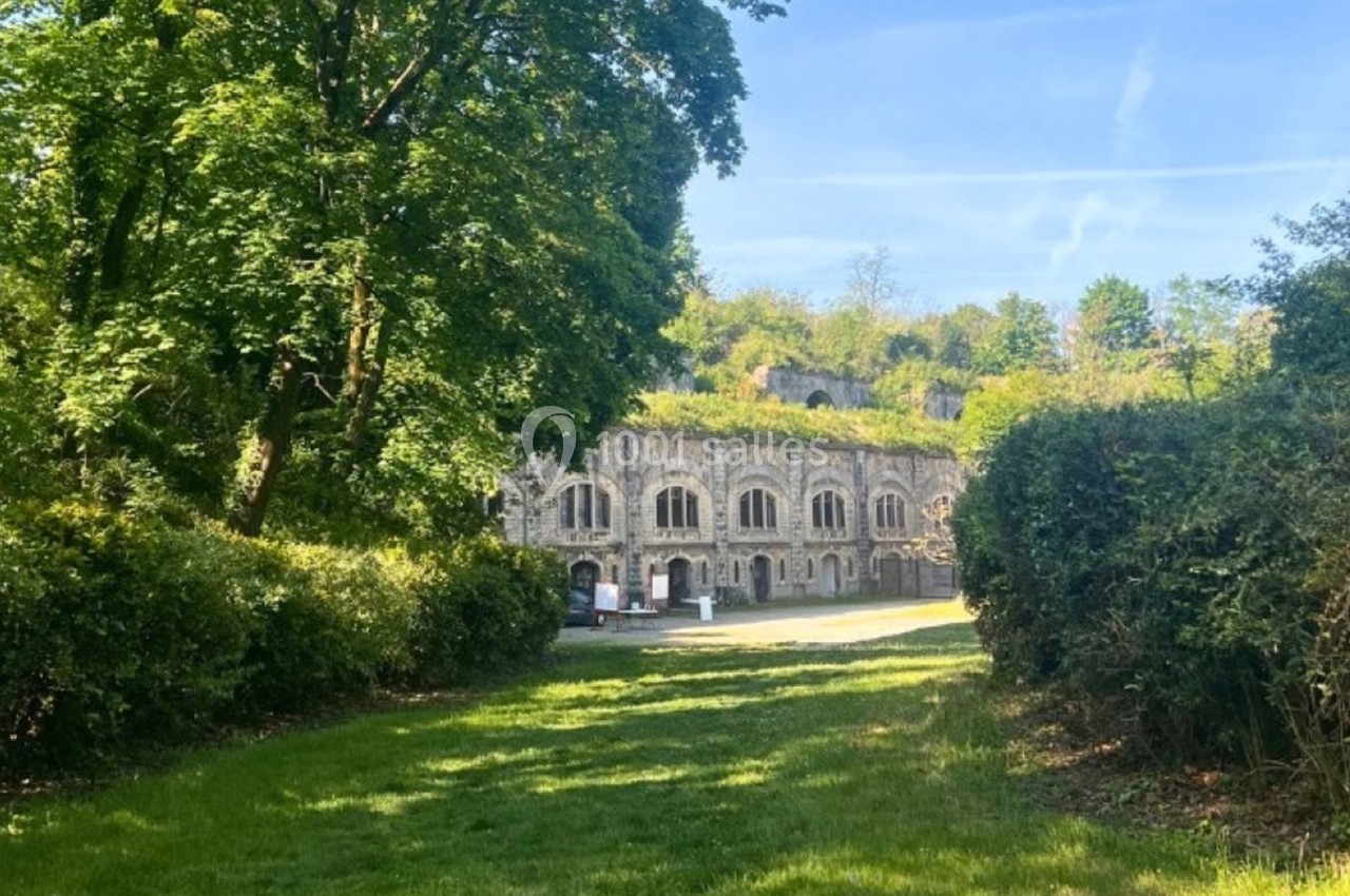 Bâtiment en pierre entouré de verdure, avec des arbres et une pelouse au premier plan sous un ciel dégagé.