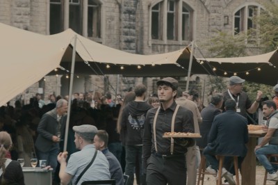 Serveur portant un plateau dans un événement en plein air sous des tentes, avec des invités assis et debout.