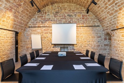 Salle de réunion en pierre avec table rectangulaire, chaises noires, rétroprojecteur, écran blanc et tableau de conférence.