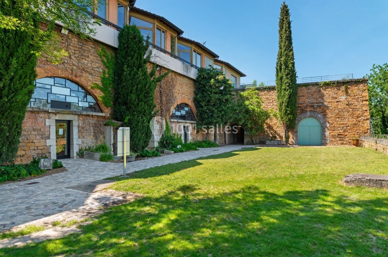 Façade en pierre d'un bâtiment avec arches, entourée de verdure et de cyprès, sous un ciel dégagé.