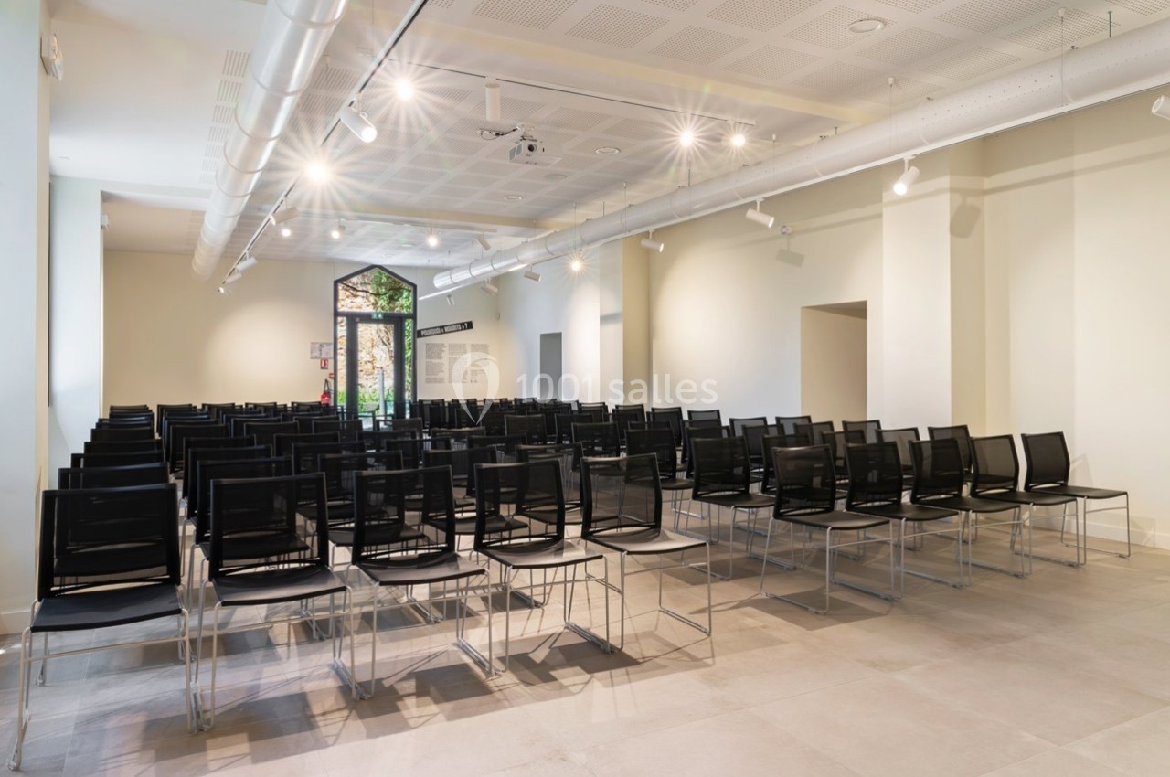 Salle de conférence lumineuse avec rangées de chaises noires alignées et une porte vitrée au fond.