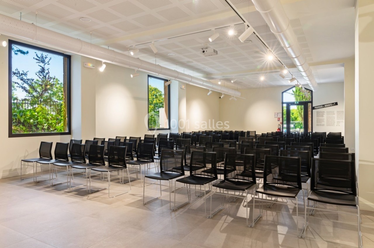 Salle lumineuse avec rangées de chaises noires alignées, grandes fenêtres donnant sur un espace vert.