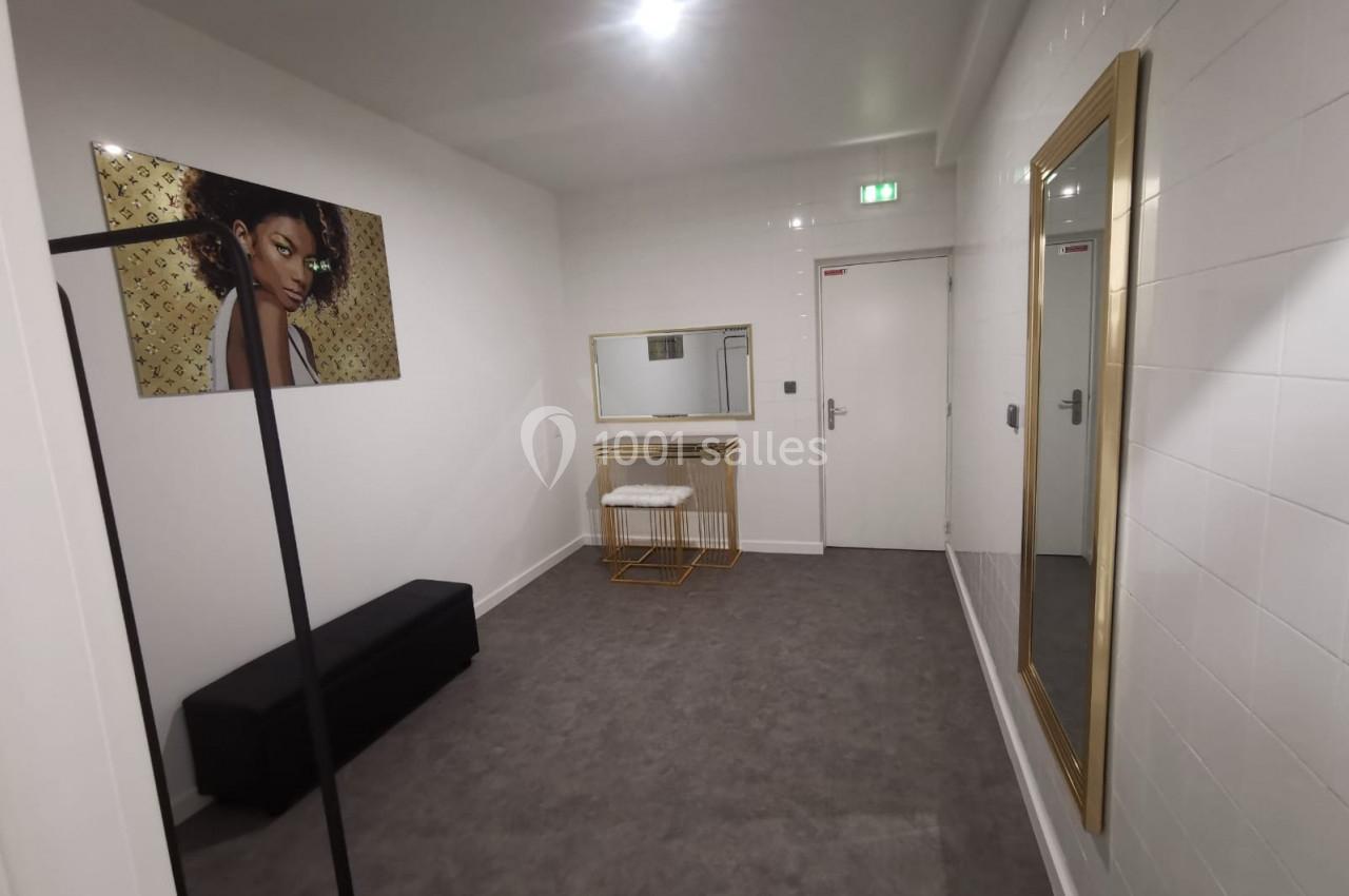 Salle lumineuse avec miroir doré, tabouret, banc noir, portant à vêtements et tableau décoratif sur un mur blanc.