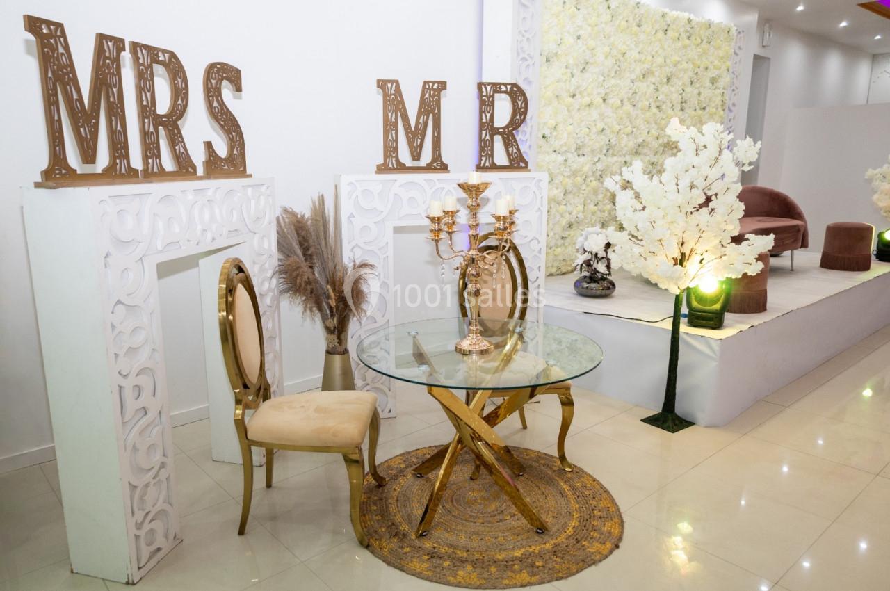 Table en verre avec chaises dorées, décorations florales blanches et inscriptions ’MRS’ et ’MR’ en arrière-plan.