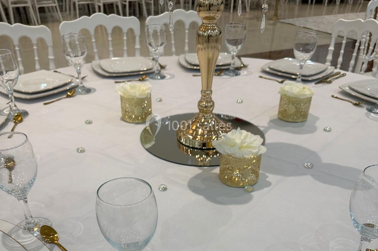 Table décorée pour un événement avec nappe blanche, bougies dorées, roses blanches et vaisselle élégante.