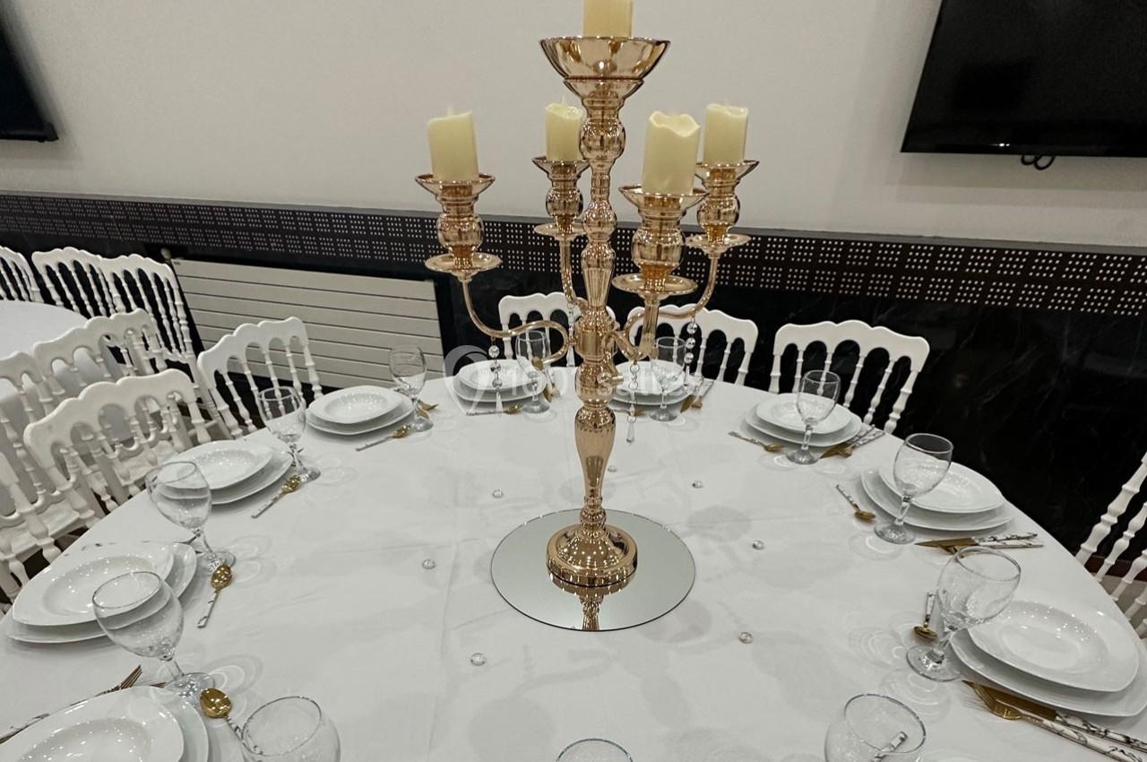 Table ronde dressée avec une nappe blanche, couverts, verres et un chandelier doré au centre avec des bougies.