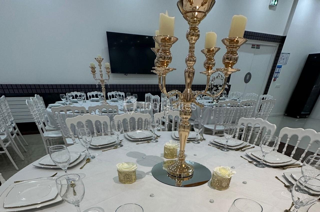 Salle de réception avec tables dressées, chandeliers dorés, vaisselle élégante et décoration florale blanche.