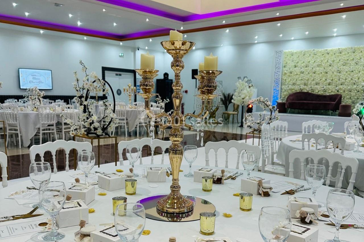 Salle de réception décorée avec des tables rondes, un chandelier doré central et des lumières d'ambiance violettes.