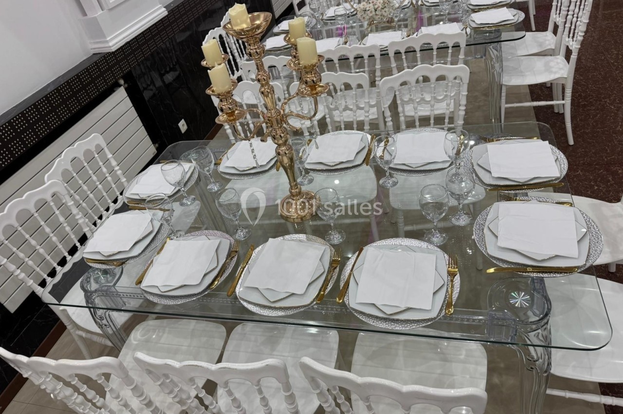 Table dressée avec vaisselle blanche, serviettes pliées, chandeliers dorés et chaises blanches dans une salle élégante.