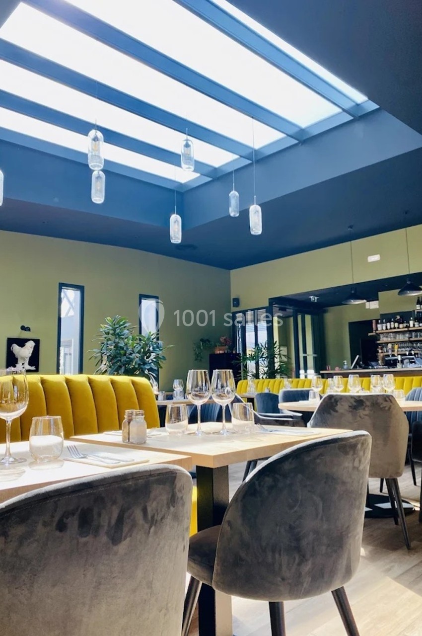 Salle de restaurant moderne avec tables dressées, banquettes jaunes, chaises grises et grande verrière laissant entrer la…