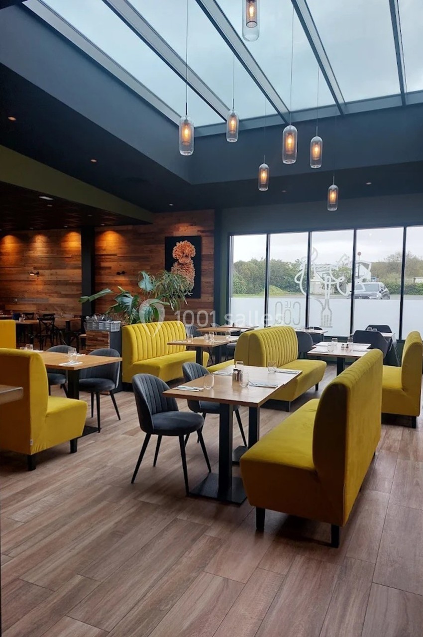 Salle de restaurant moderne avec banquettes jaunes, tables en bois, grandes baies vitrées et plafond vitré.