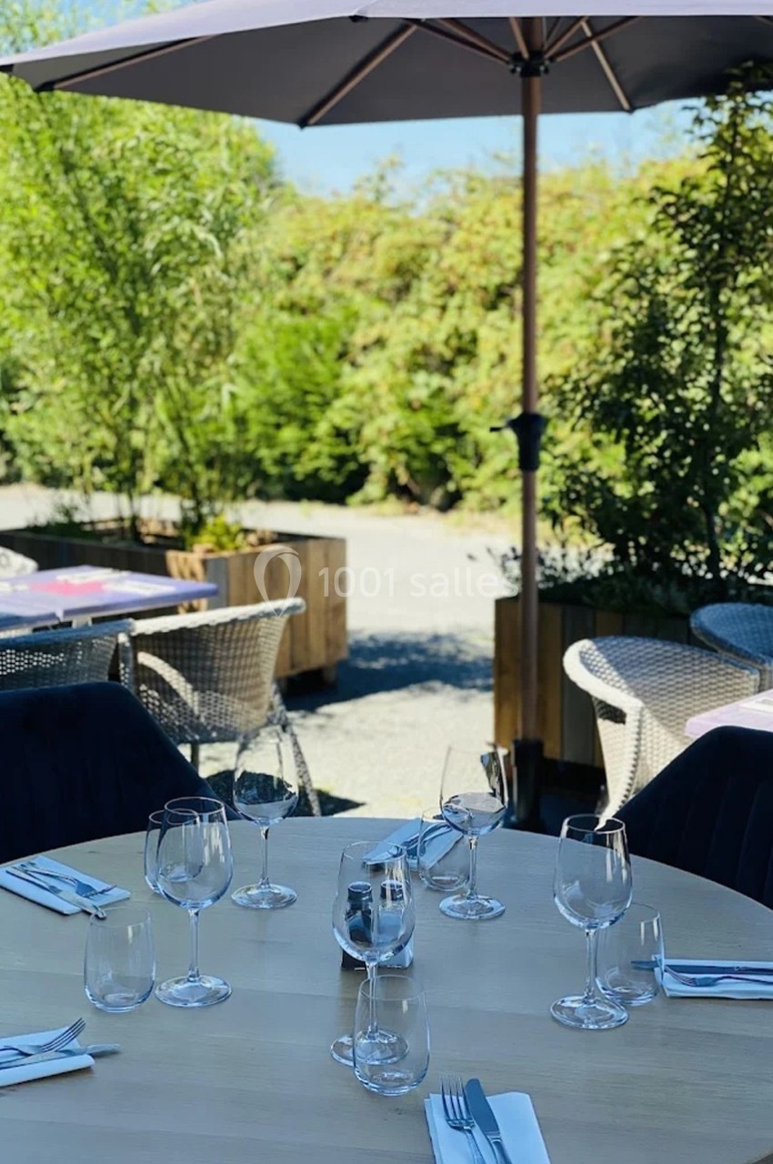 Table dressée en extérieur avec verres et couverts, entourée de chaises, sous un parasol et près de végétation.