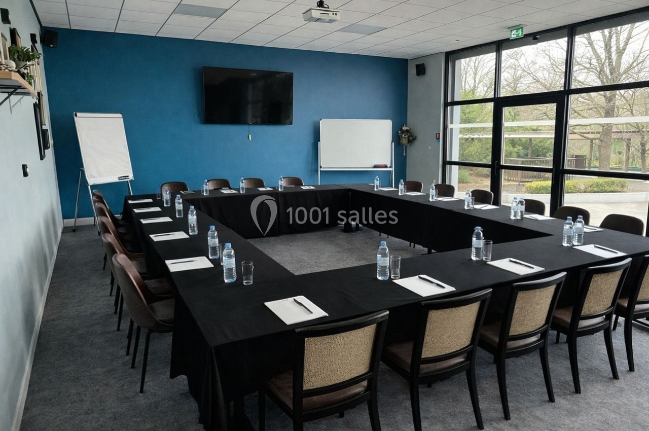 Salle de réunion avec tables disposées en U, chaises, écran, paperboard et vue sur l'extérieur par des baies vitrées.