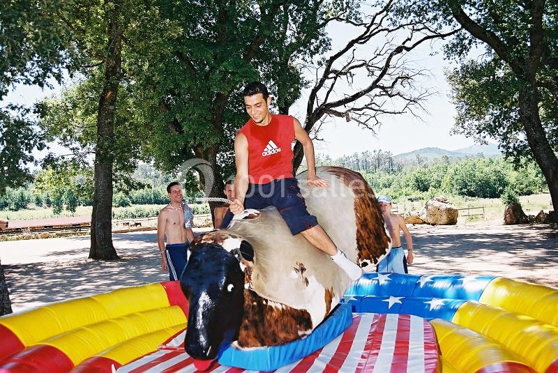 Un homme monte un taureau mécanique dans un espace extérieur entouré d'arbres, sous un ciel dégagé.