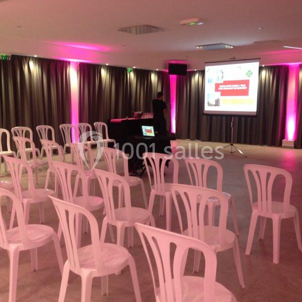 Salle de conférence avec des chaises blanches alignées, un écran de projection et un éclairage rose.