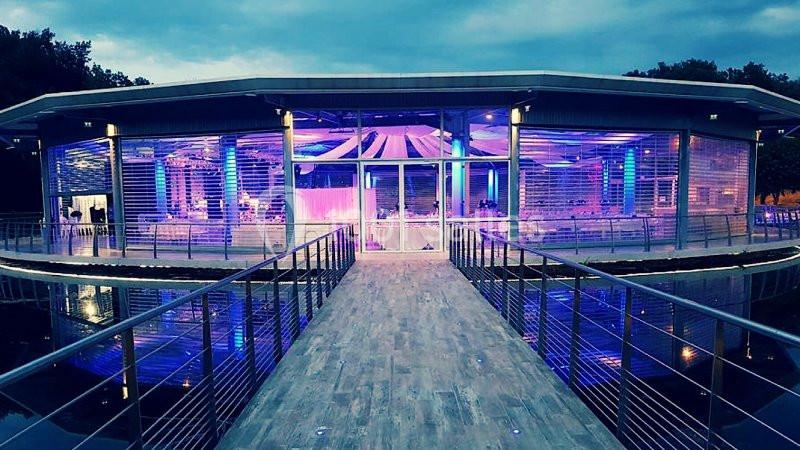 Salle moderne éclairée en bleu, entourée de baies vitrées, accessible par une passerelle au-dessus de l'eau.