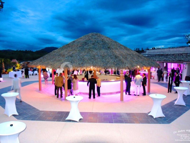 Personnes rassemblées autour d'un bar circulaire sous une paillote éclairée, dans un espace extérieur en soirée.