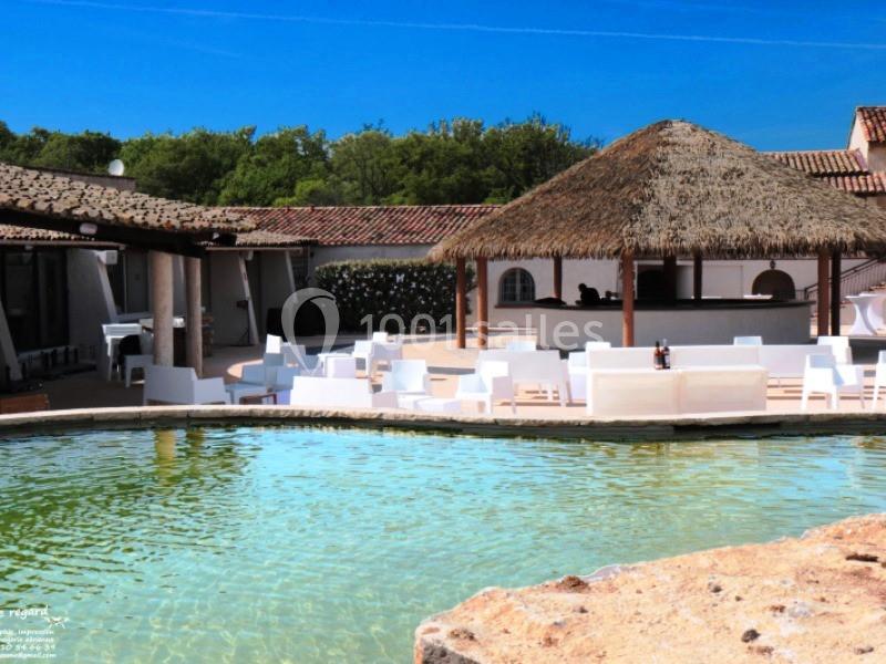 Espace extérieur avec piscine, mobilier blanc, bar au toit de chaume et bâtiments entourés de végétation.