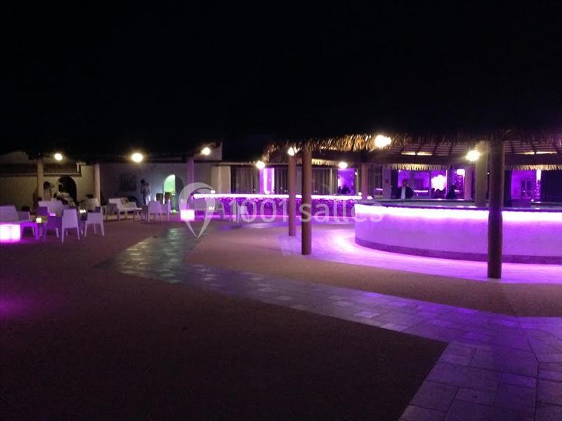 Bar extérieur éclairé par des lumières violettes, avec des tables et des chaises disposées sur une terrasse de nuit.