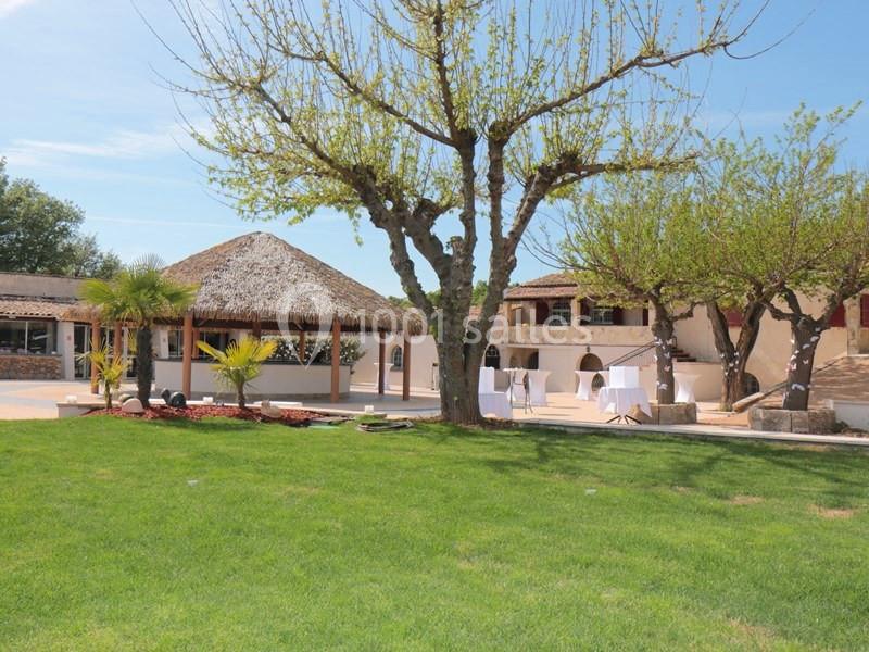 Cour extérieure avec pelouse, arbres, terrasse ombragée et bâtiments en pierre sous un ciel dégagé.