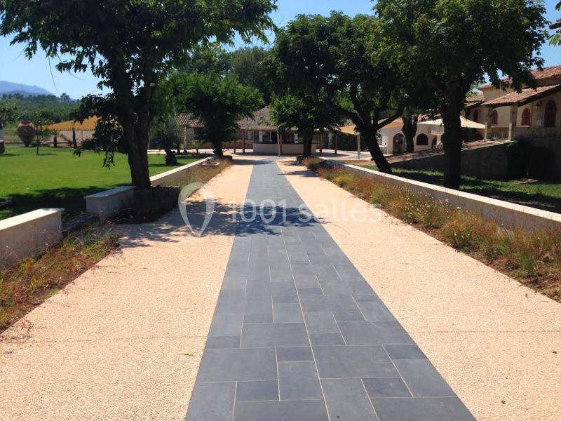 Allée pavée bordée d'arbres et de végétation, menant à des bâtiments dans un cadre ensoleillé.