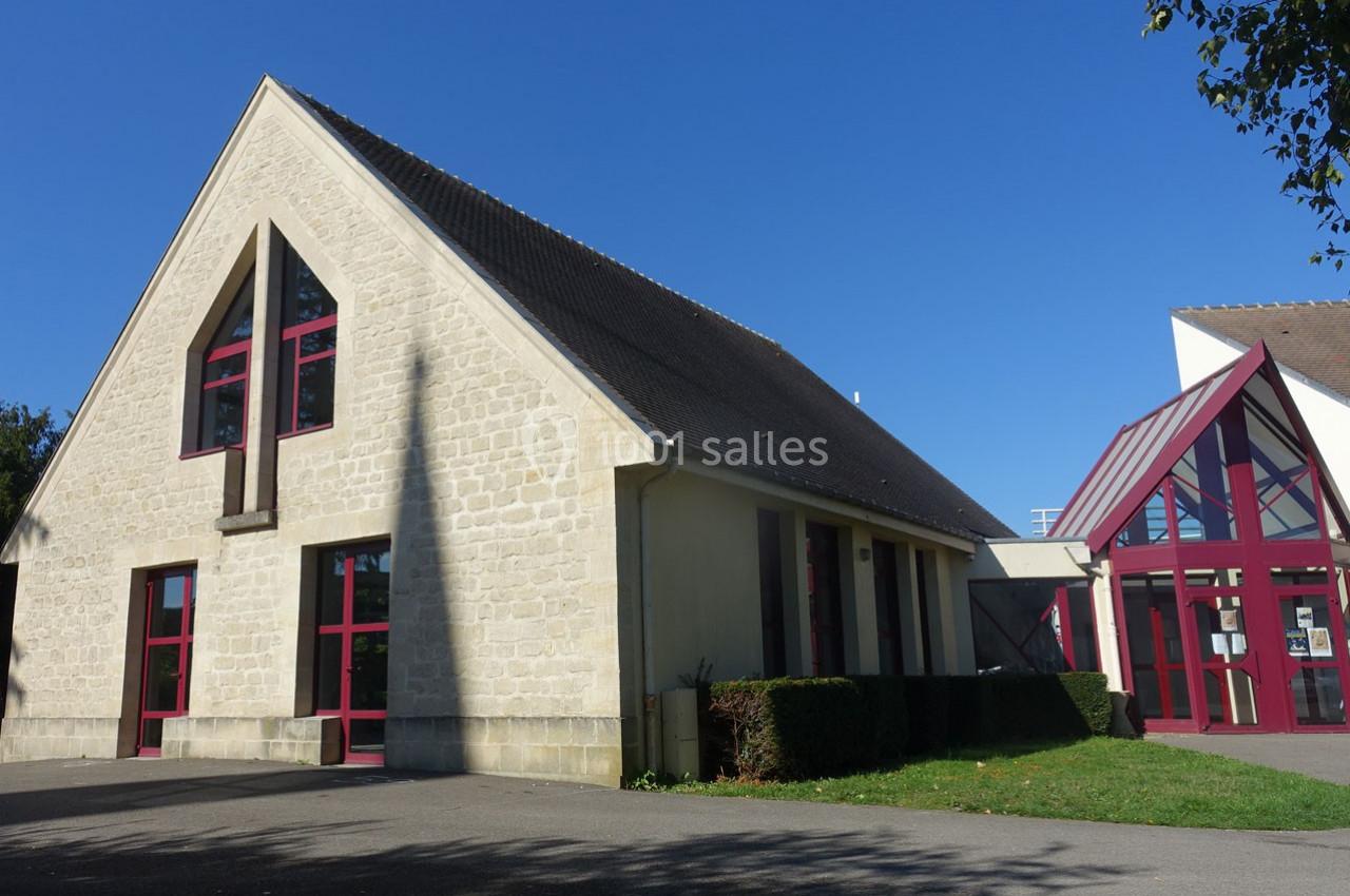 Bâtiment en pierre claire avec toit en ardoise, grandes fenêtres rouges et entrée vitrée sous un ciel bleu.