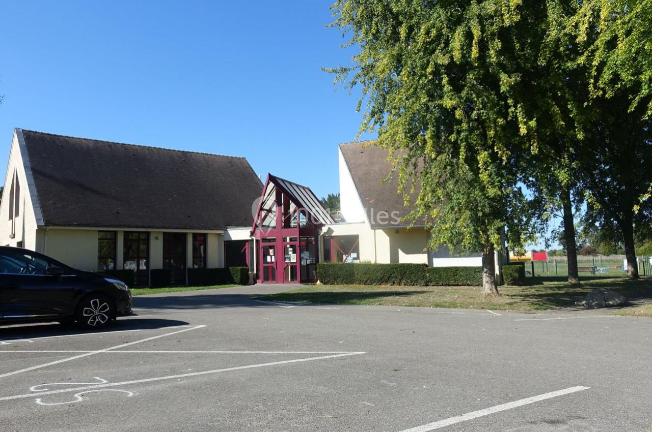 Bâtiment à toit en pente avec entrée vitrée, entouré d'arbres, vu depuis un parking ensoleillé.