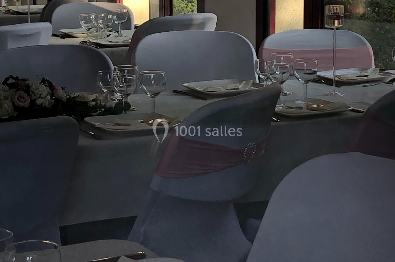 Salle de réception avec tables dressées, nappes blanches, chaises décorées et lumière naturelle entrant par les fenêtres.