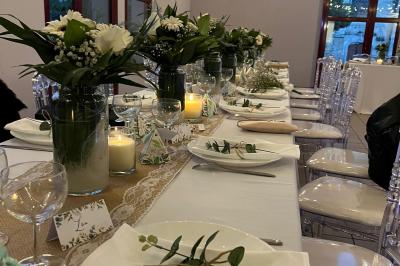 Salle de réception avec tables dressées, nappes blanches, chaises décorées et lumière naturelle entrant par les fenêtres.