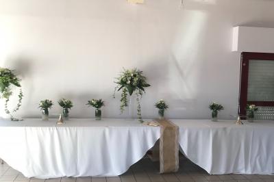 Salle de réception avec tables dressées, nappes blanches, chaises décorées et lumière naturelle entrant par les fenêtres.