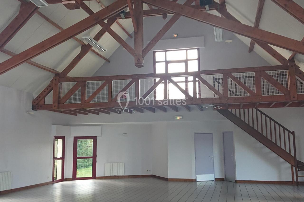 Salle spacieuse avec charpente en bois apparente, carrelage au sol, mezzanine et grandes fenêtres laissant entrer la lumière.
