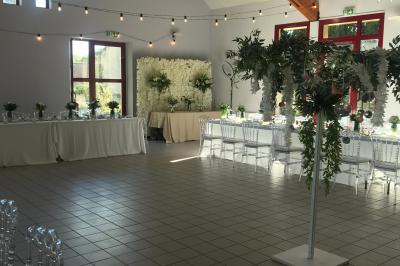 Salle de réception avec tables dressées, nappes blanches, chaises décorées et lumière naturelle entrant par les fenêtres.