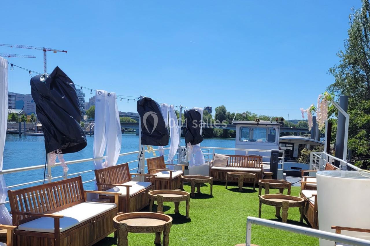 Terrasse d'un bateau aménagée avec des fauteuils en bois, des tables rondes et des voilages blancs, sur fond de rivière.
