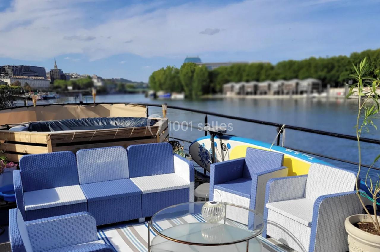 Mobilier de jardin bleu et blanc sur une terrasse au bord d'une rivière, avec jacuzzi et végétation en arrière-plan.