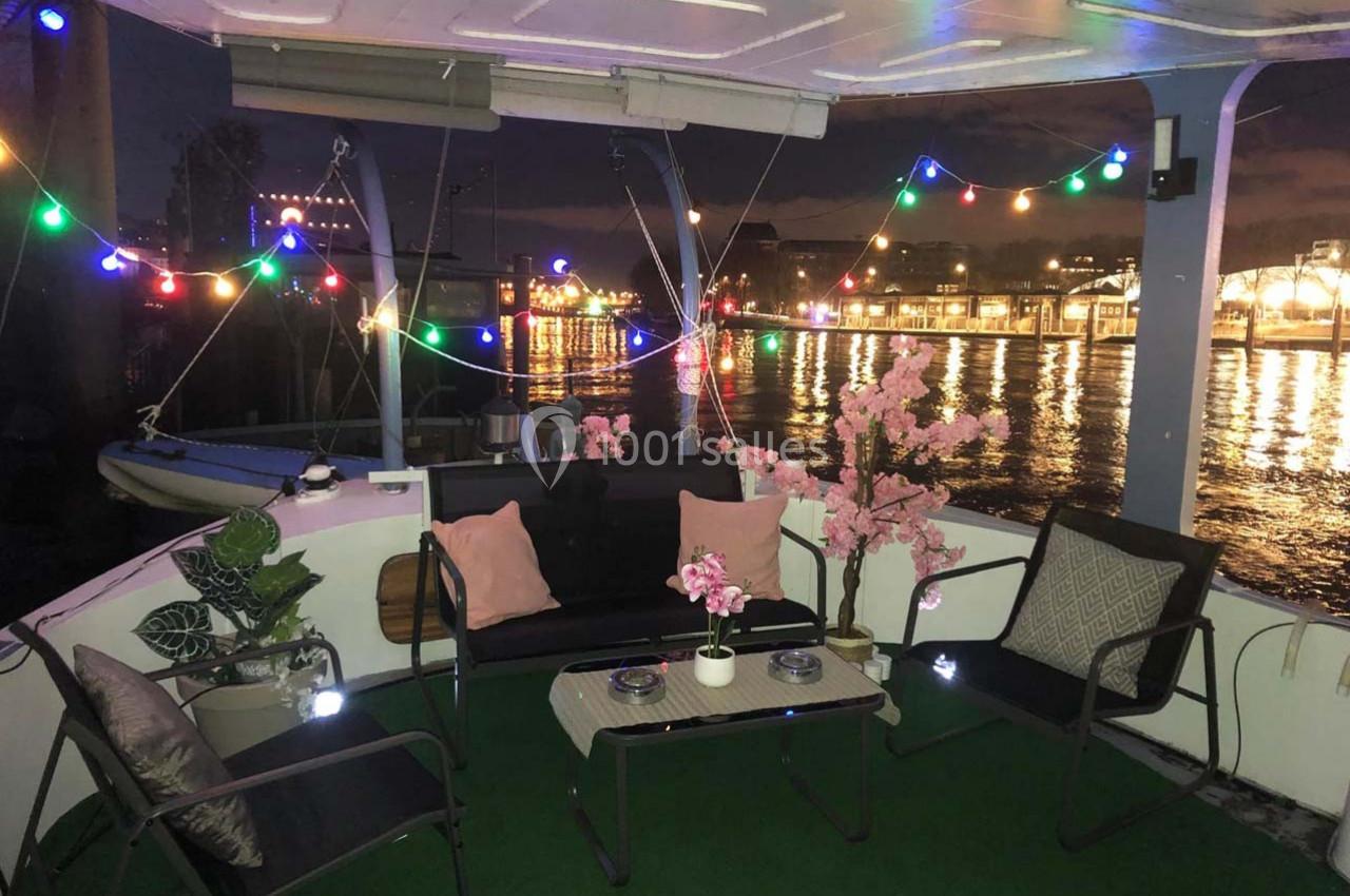 Espace extérieur d'un bateau aménagé avec des meubles, des plantes et des guirlandes lumineuses, vue sur un port éclairé.