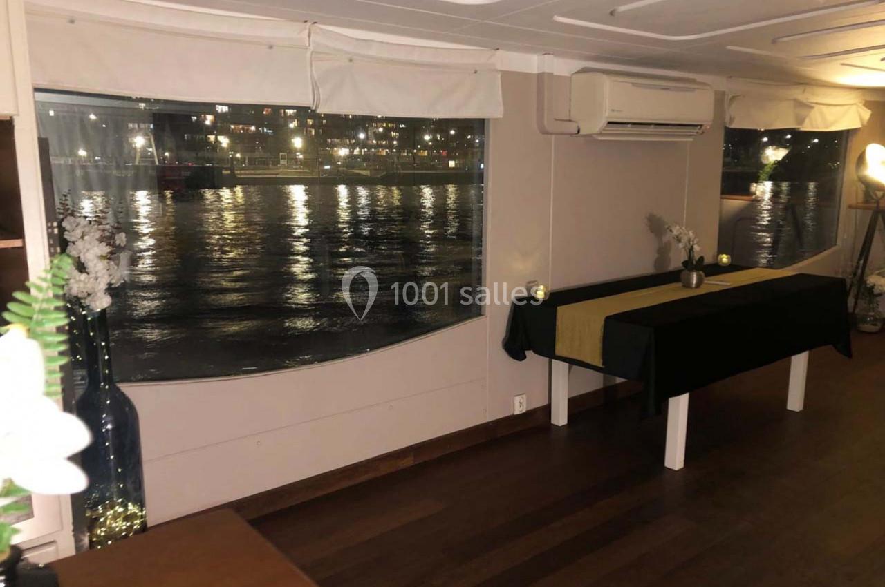 Intérieur d'une salle avec vue sur une rivière illuminée la nuit, tables décorées et éclairage tamisé.