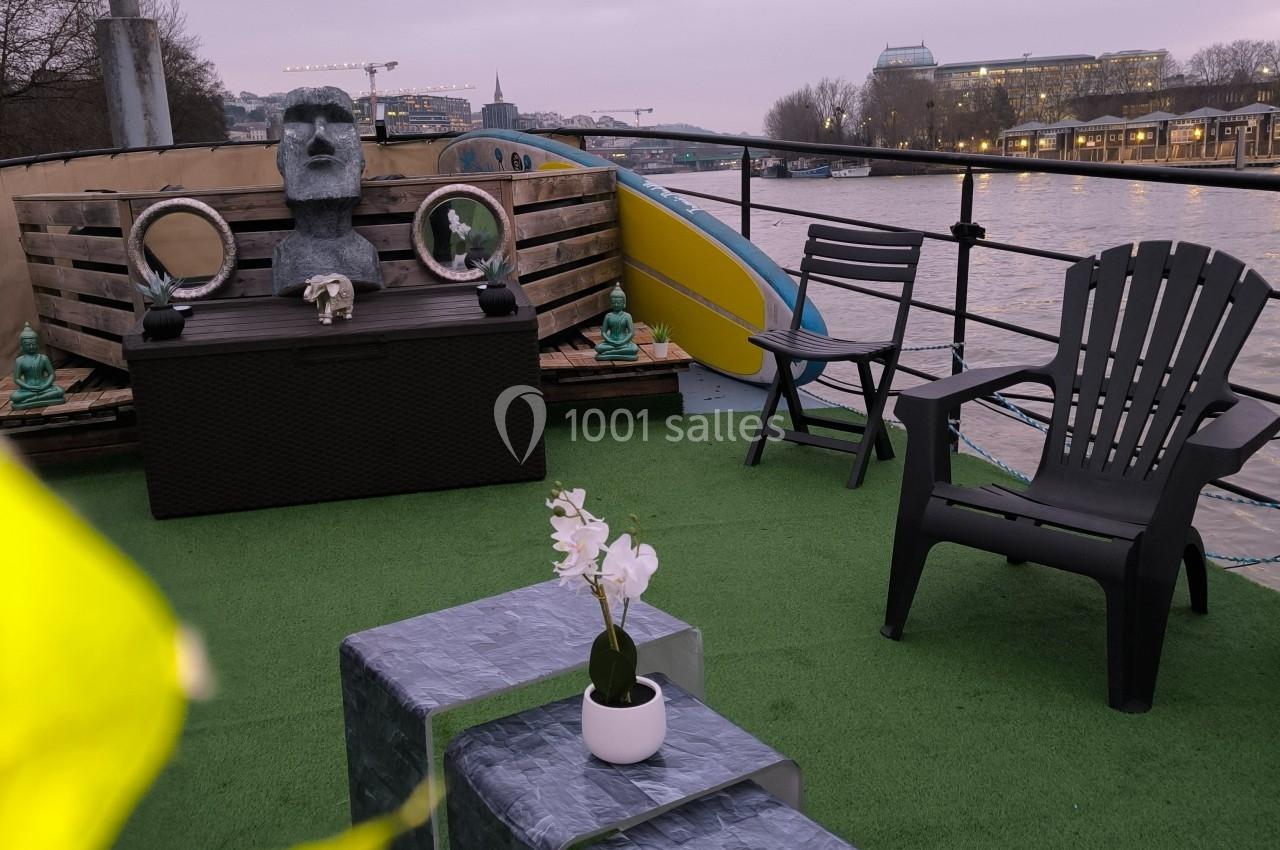 Terrasse aménagée sur une péniche avec fauteuils, table basse, orchidée, statue et vue sur un fleuve au crépuscule.