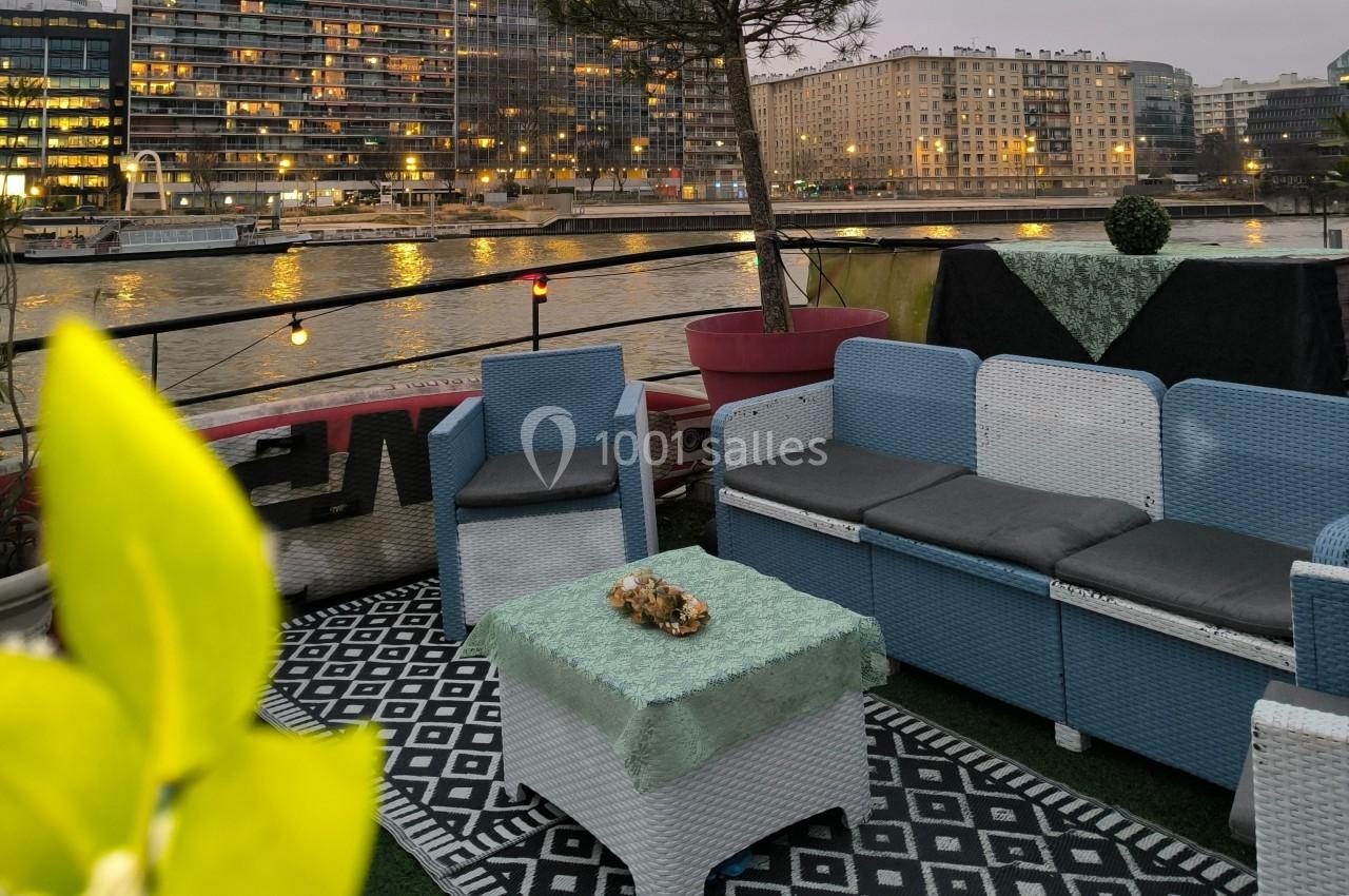 Mobilier de jardin en rotin avec coussins, installé sur une terrasse au bord de l'eau, entouré de bâtiments illuminés.