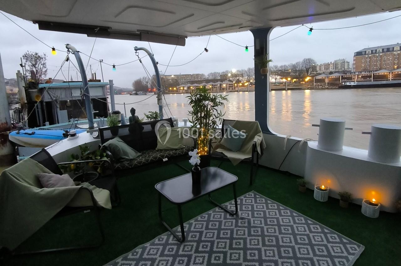Salon extérieur aménagé sur une péniche, avec mobilier confortable, plantes et vue sur une rivière bordée de bâtiments.